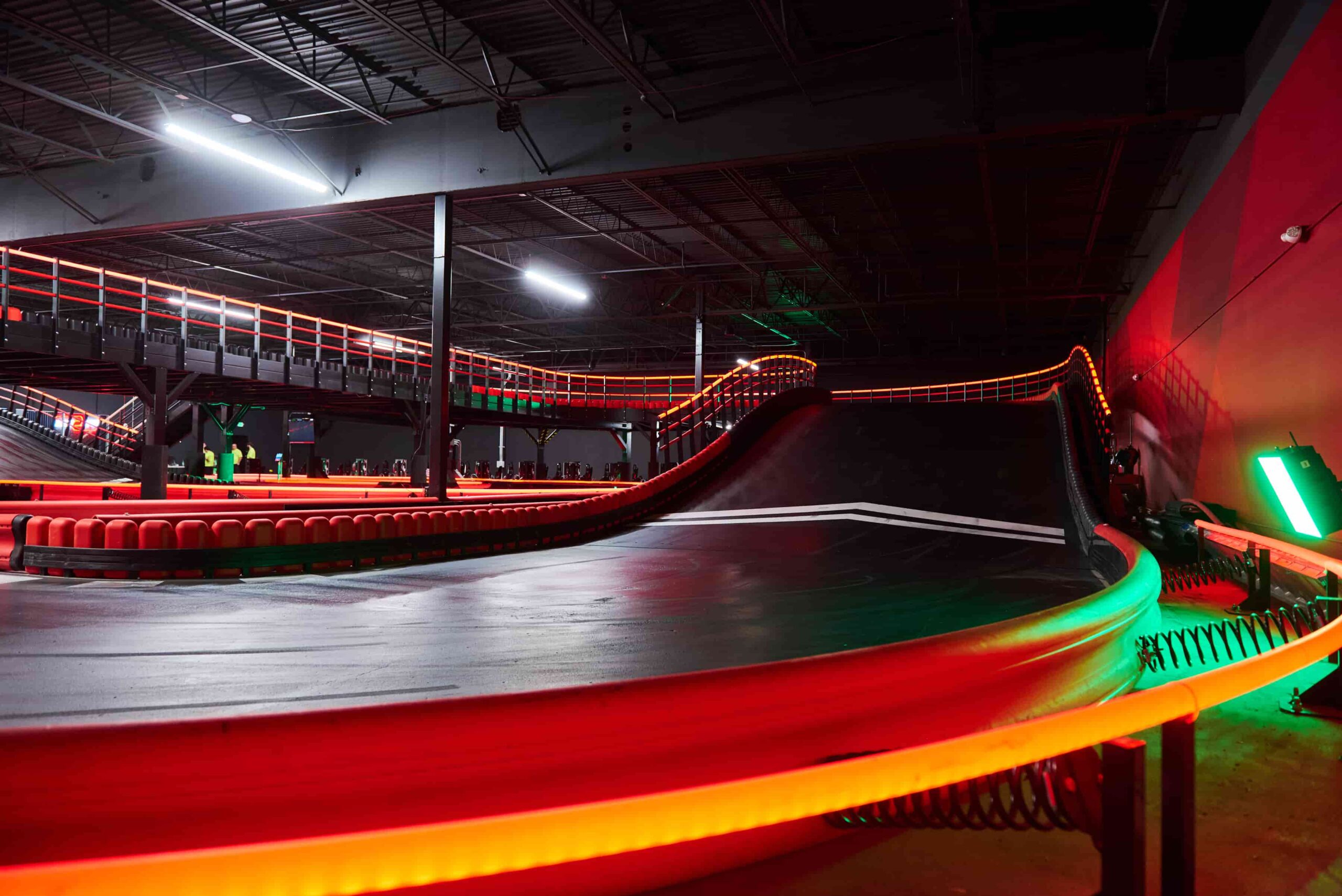 An indoor go-kart track with a ramp, elevated circuit, and red safety barriers curves around a tall wall.