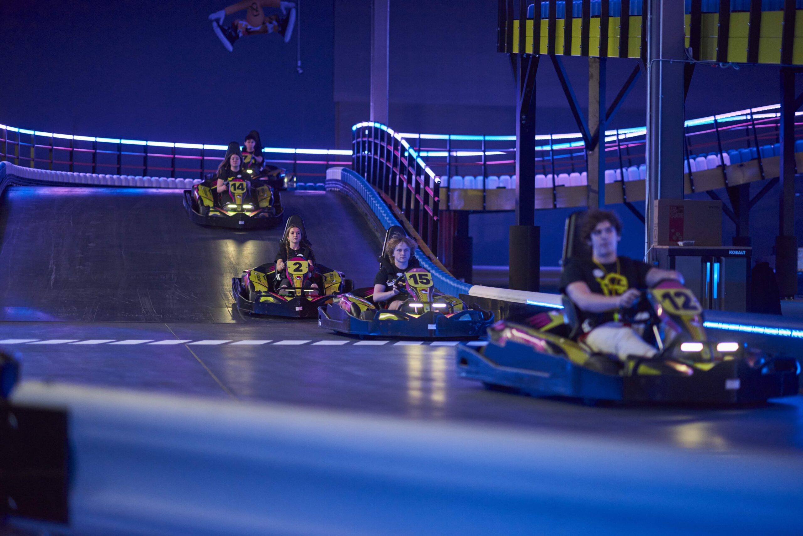 Several people drive go-karts around an indoor track with blue lighting, guardrails, and a checkered floor marking.